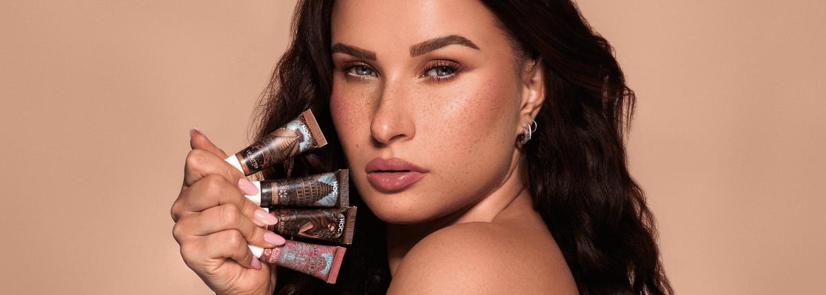 Model mit gepflegten, glänzenden Lippen hält alle 4 NOC. Lip Butter mit Peptiden: Occult Glow, Sin Tower, Hedon Flame und Lust Garden