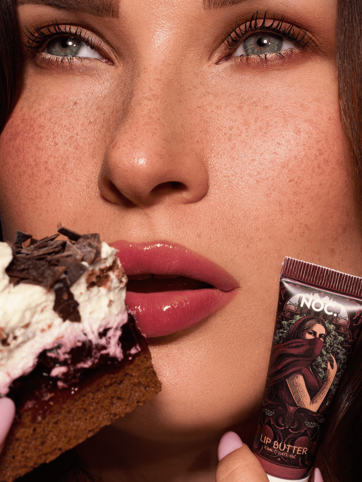 Model mit glänzenden Lippen hält Hedon Flame Lip Butter von NOC. Cosmetics neben einem Stück Kirschtorte