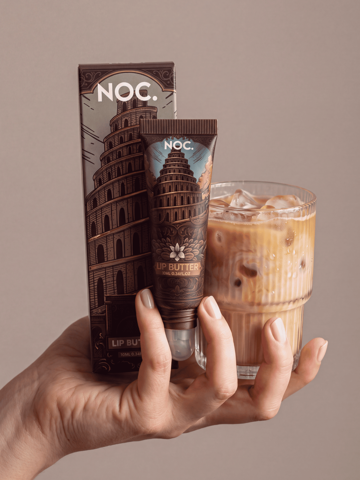 Hand hält Sin Tower Lip Butter von NOC. Cosmetics mit Verpackung vor einem Iced Coffee, getönter Lippenbalsam mit Kaffee-Aroma